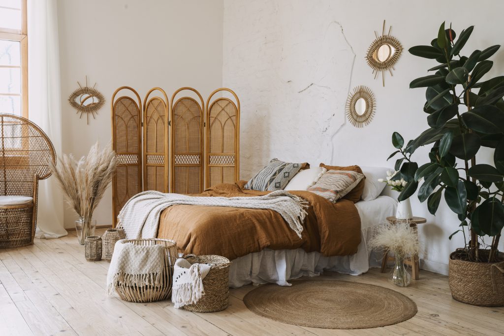Comfortable apartment in bohemian style interior with hygge bedroom, pillow and bedspread on bed, bamboo dressing screen, home decor, dry plants in vase, wicker basket, houseplant on floor